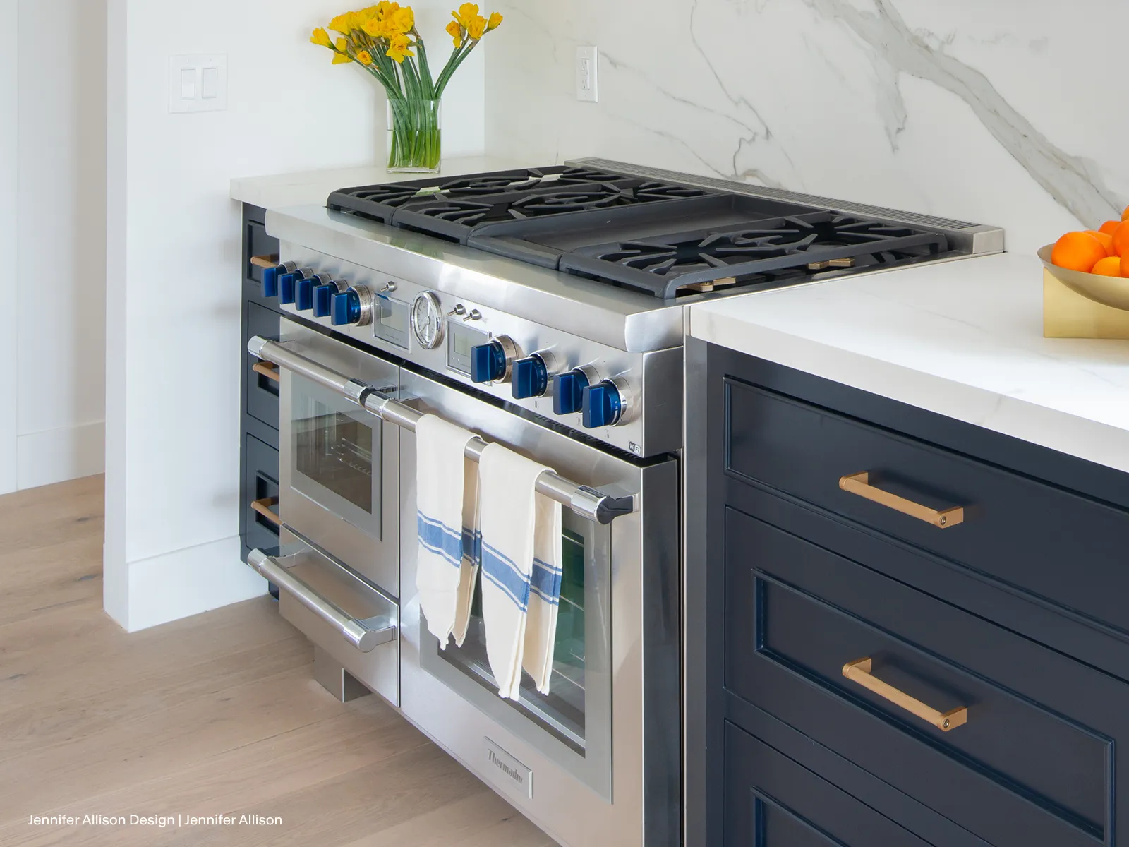 Modern stainless steel gas stove with blue knobs, navy cabinets, white marble countertop, yellow flowers, and a bowl of oranges.