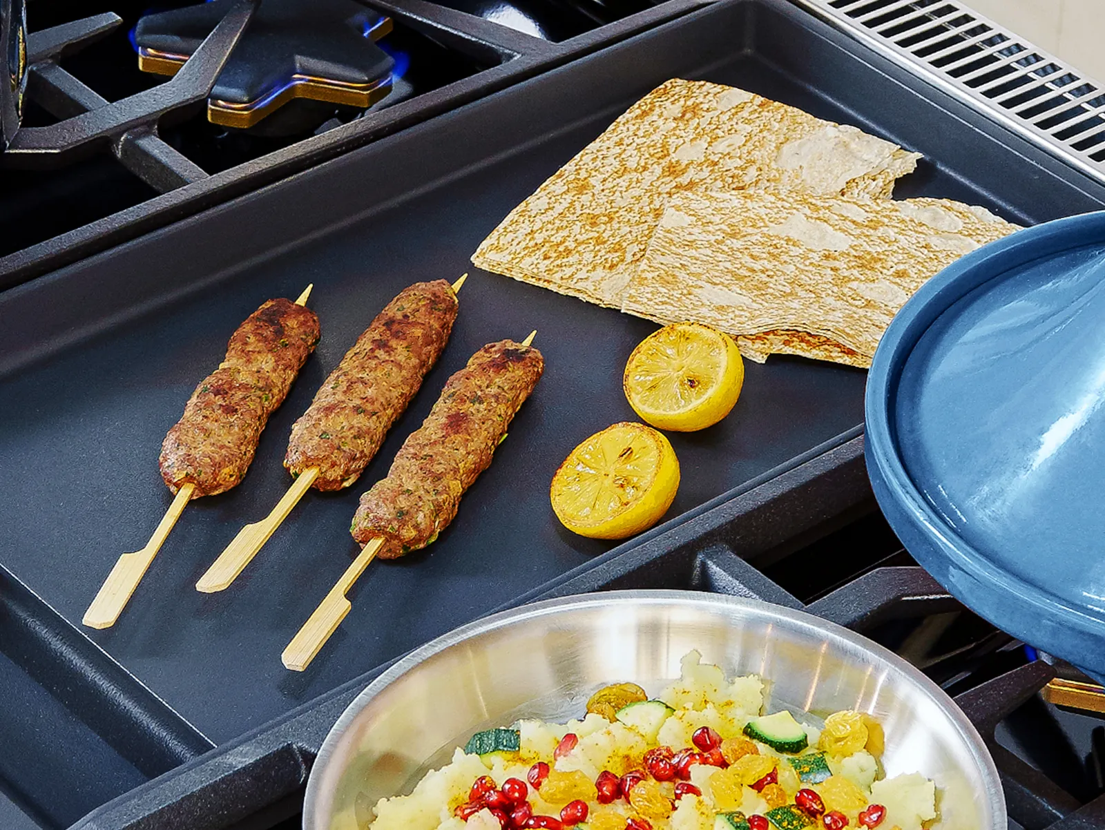 Three grilled meat skewers, flatbreads, and halved grilled lemons on a griddle next to a pan with colorful cooked vegetables.