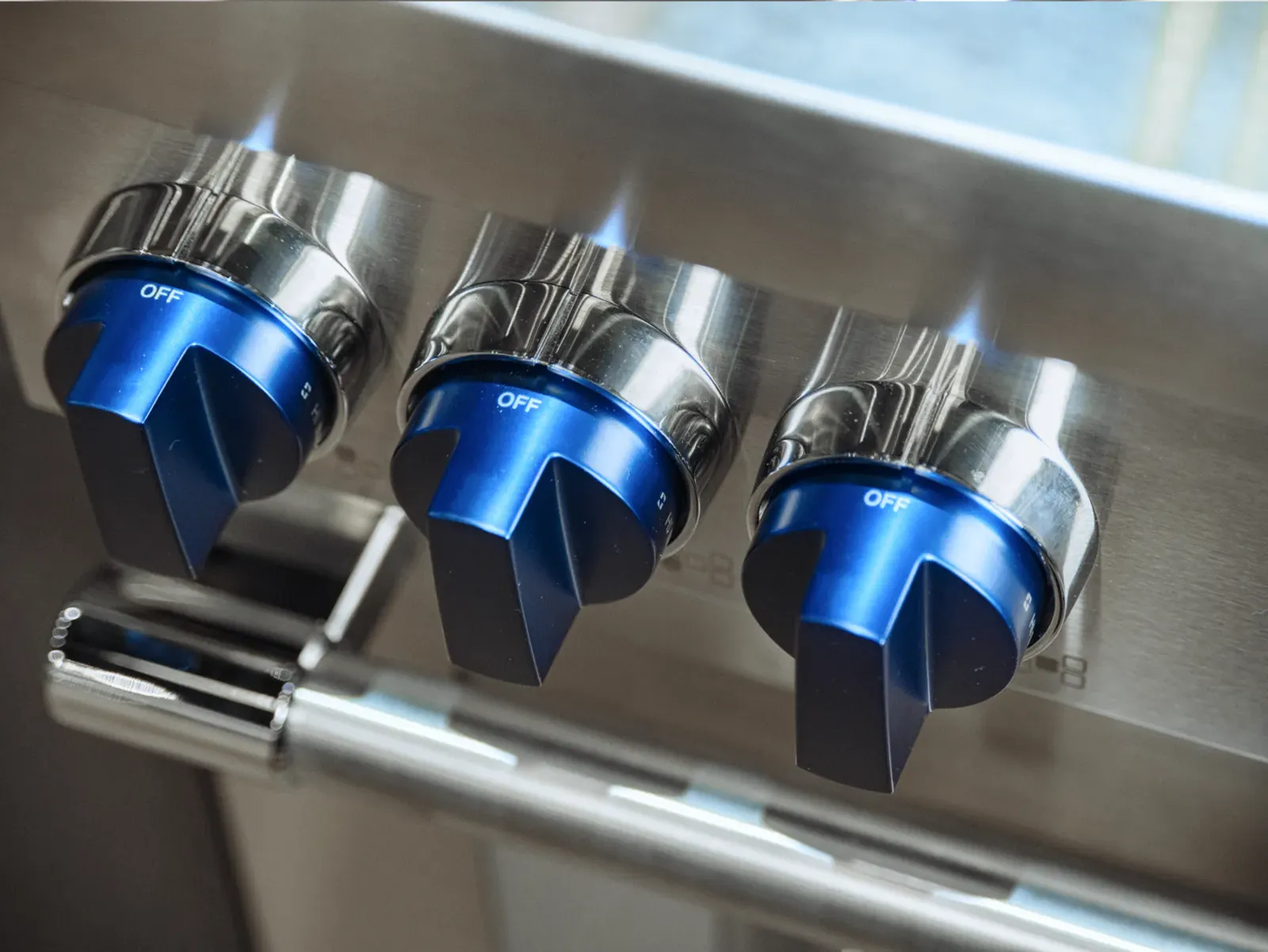 A close-up, angled shot of three signature Thermador blue metallic control knobs on a stainless steel range, all set to the "OFF" position.