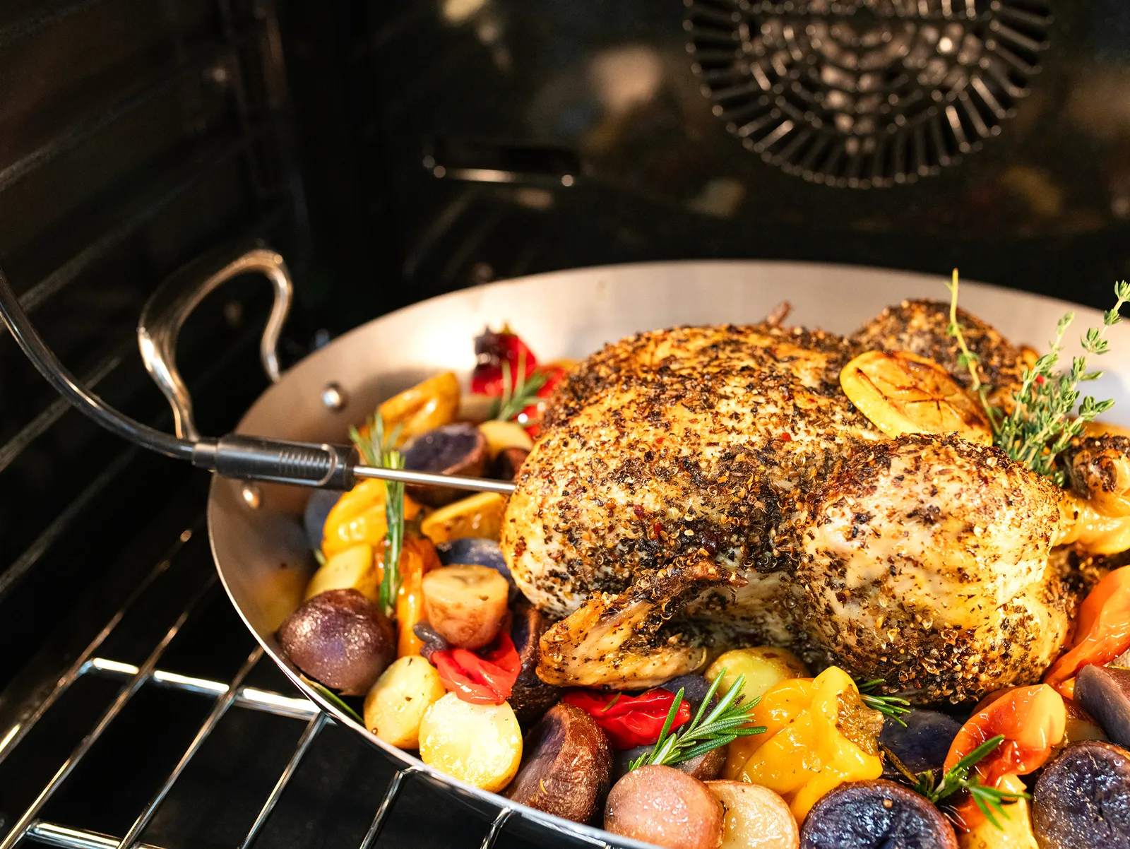 Whole seasoned chicken roasting in an oven, surrounded by multicolored potatoes, bell peppers, and fresh herbs in a large pan.