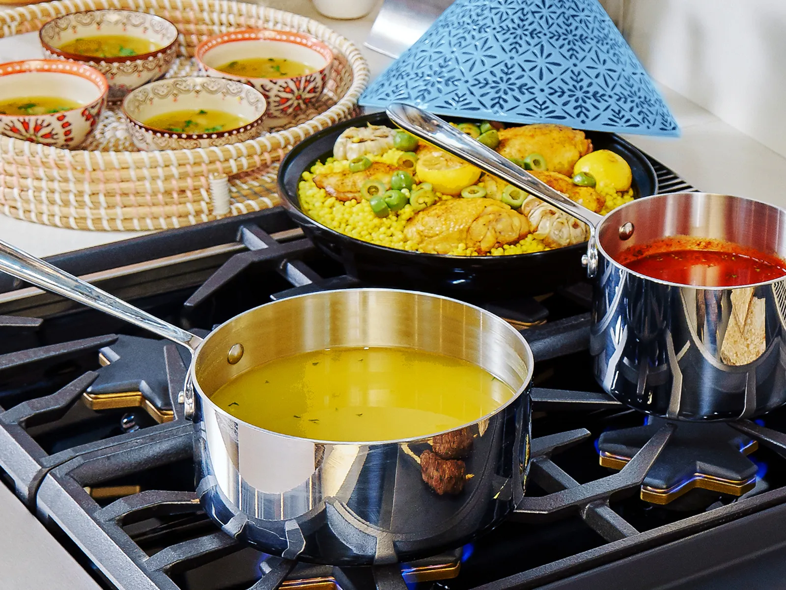 Two saucepans with yellow and red sauces on a gas stove, a pan with yellow rice and chicken, and a tray with bowls of soup in the background.