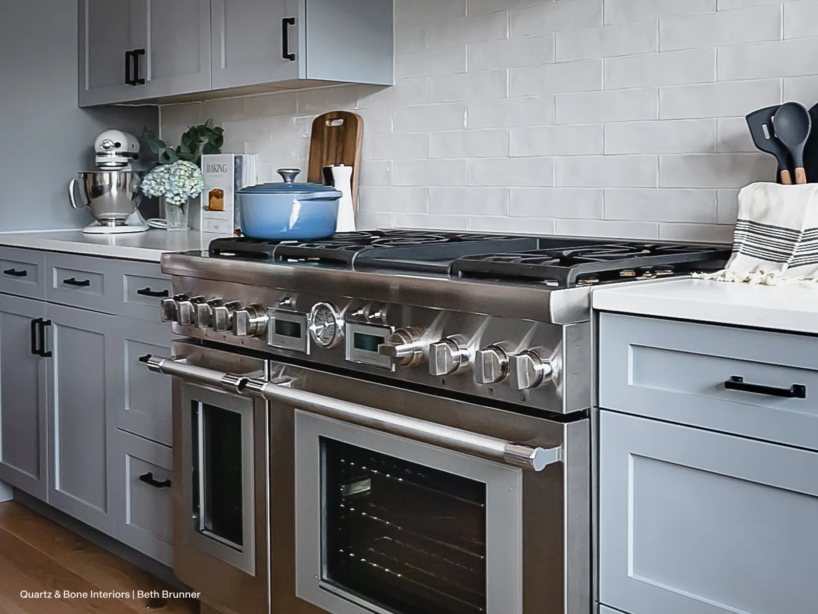 A stainless steel professional-style range is the centerpiece of a modern kitchen featuring light gray shaker cabinets and white countertops. The double oven range has a blue Dutch oven sitting on its gas cooktop, complementing the kitchen's clean and stylish aesthetic.