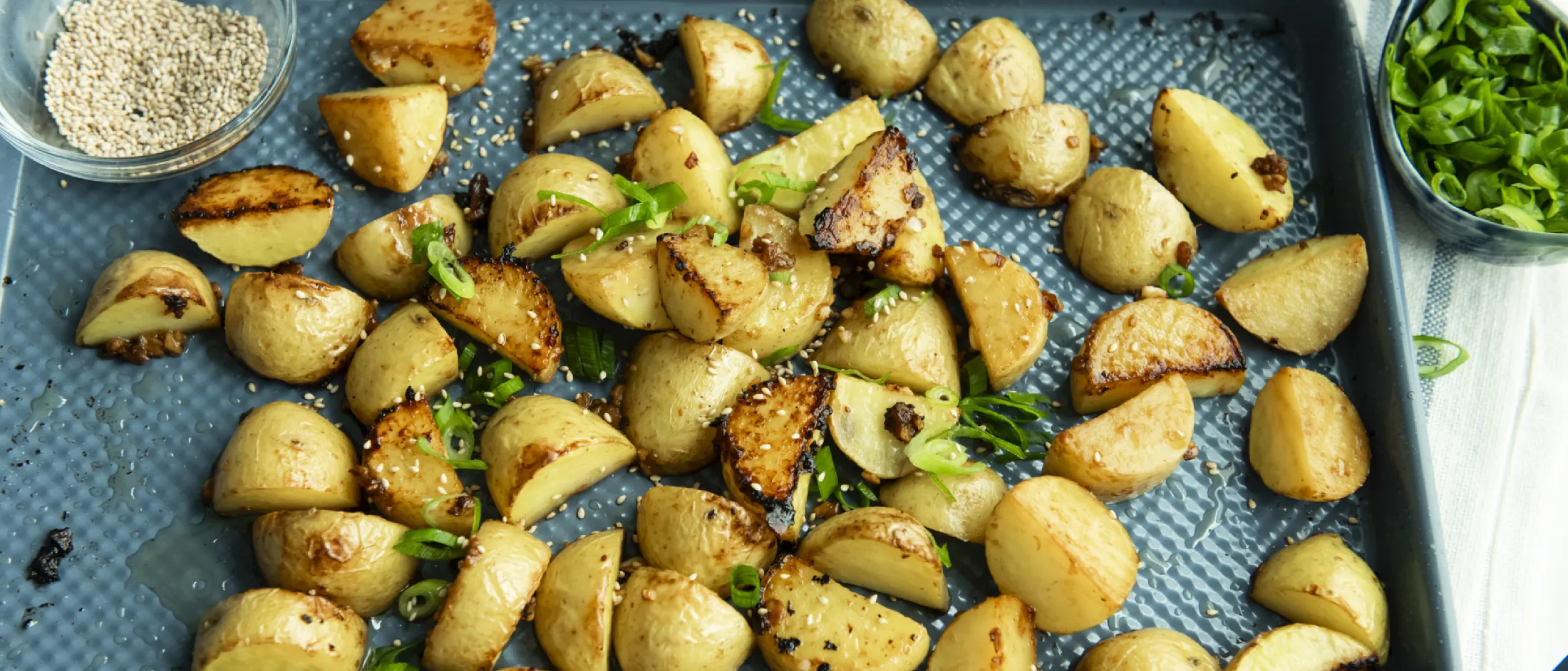 Tray of roasted potato halves garnished with chopped green onions and sesame seeds with bowls of sesame seeds and chopped green onions on the side