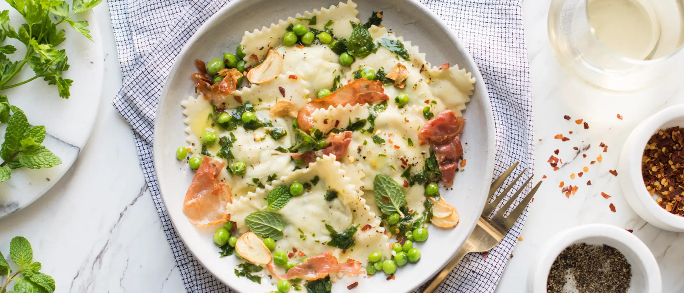Plate of ravioli with peas, herbs, garlic, and prosciutto on a cloth napkin, with pepper, chili flakes, and a glass of white wine nearby.