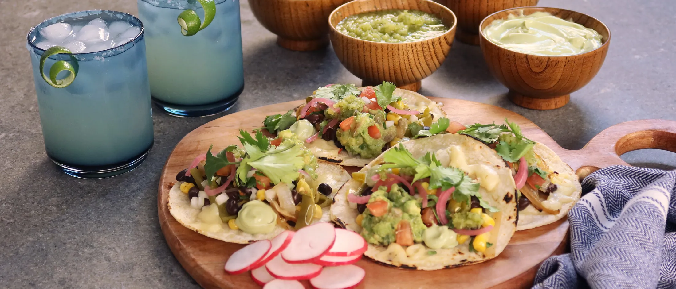 Four veggie tacos topped with guacamole and cilantro on a wooden board with sliced radishes, sauces, and two margaritas.