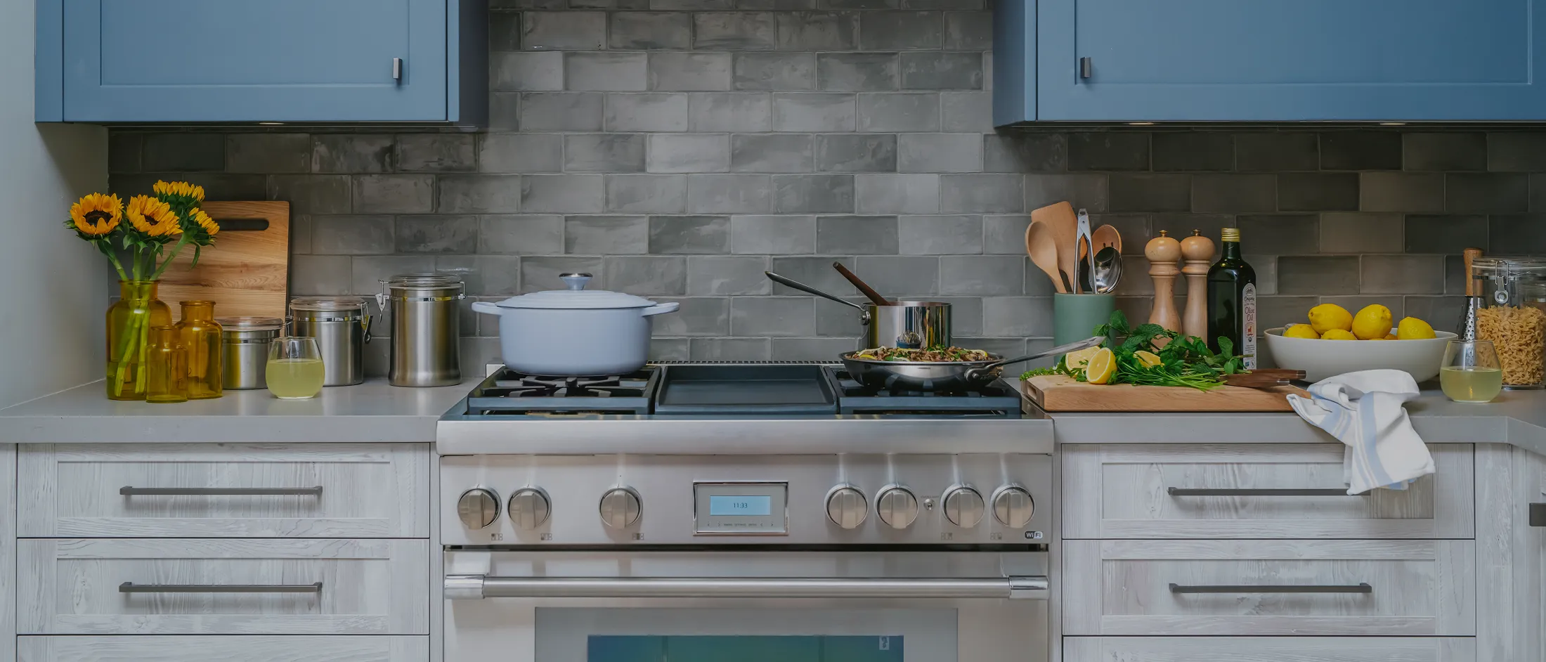 A stainless steel range is centered in a stylish kitchen, flanked by light wood-grain lower cabinets and set against a dark gray tile backsplash. Dusty blue upper cabinets provide a colorful contrast. The countertops are styled with fresh ingredients, including a vase of sunflowers and a bowl of lemons, while a light blue Dutch oven and a frying pan sit on the cooktop.