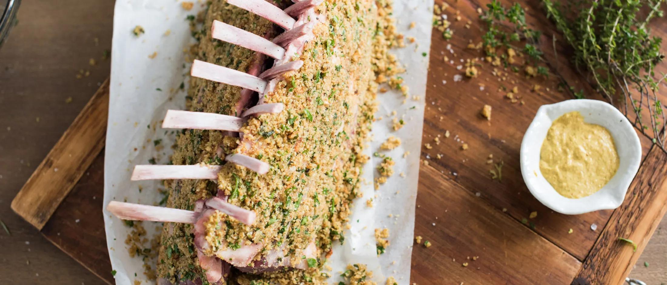 Sliced meat on a wooden board, surrounded by herbs, spices, and a small bowl of sauce.
