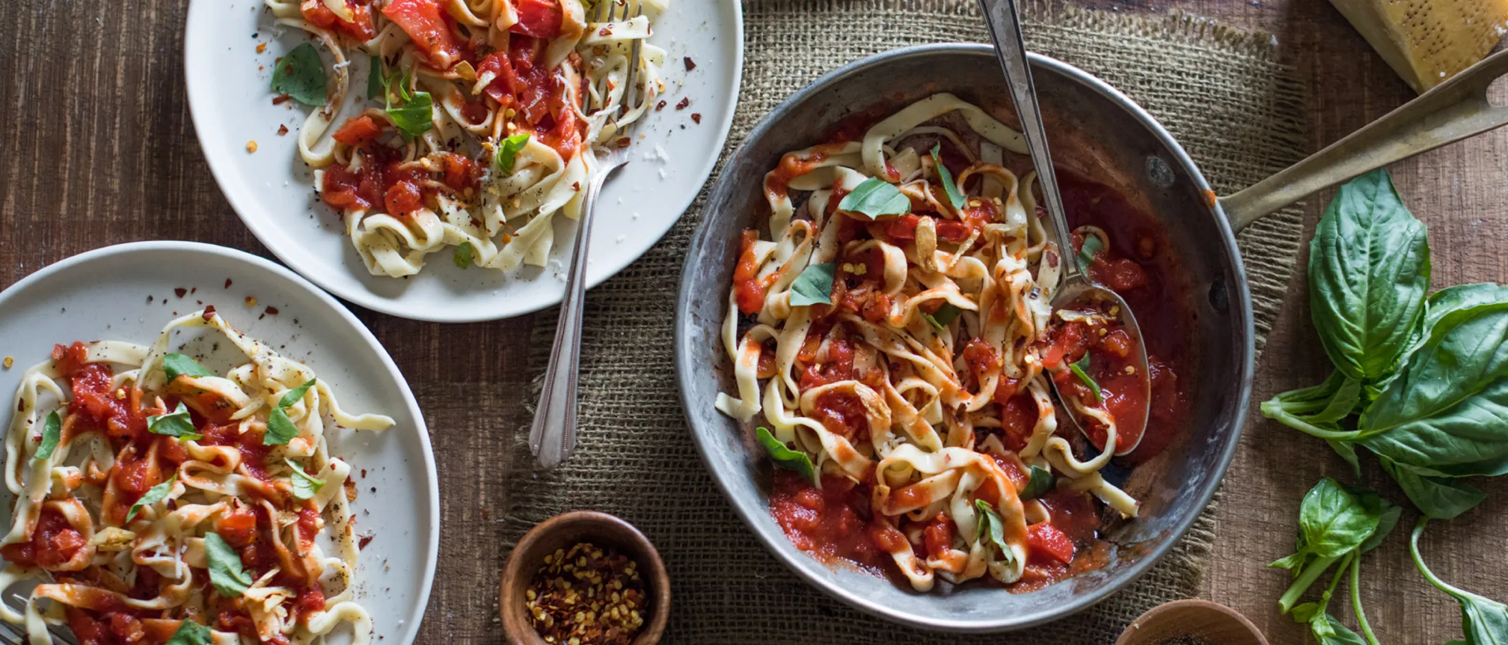 Two plates and a pan of pasta with tomato sauce and basil, next to bowls of pepper, chili flakes, fresh basil, and a block of cheese.