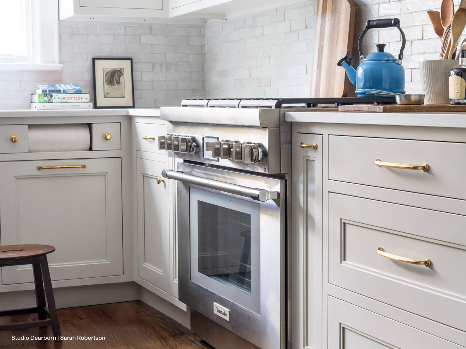 A stainless steel professional-style range is nestled in a bright kitchen corner between off-white shaker cabinets accented with gold hardware. A bright blue kettle sits on the gas cooktop, contrasting with the light-colored brick backsplash and dark hardwood floors.