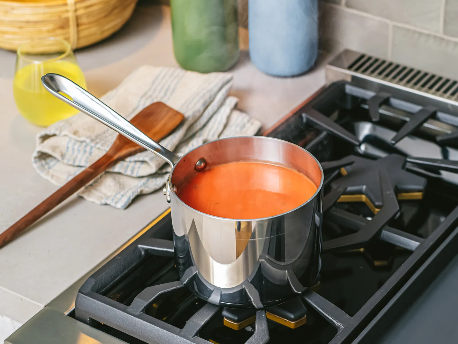 A close-up shot of a stainless steel saucepan filled with simmering tomato soup, sitting on a distinctive black, star-shaped gas burner. Steam rises from the pot, and a wooden spoon and a glass of juice are visible in the background on the countertop.