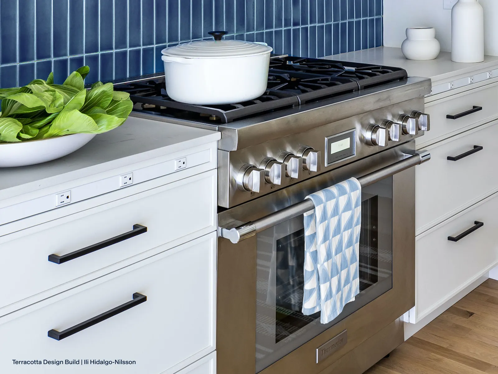 A stainless steel professional-style range is set against a vibrant, vertically stacked blue tile backsplash in a modern kitchen. The range is flanked by white cabinets with contrasting black hardware, and a blue and white geometric towel hangs from the oven handle.