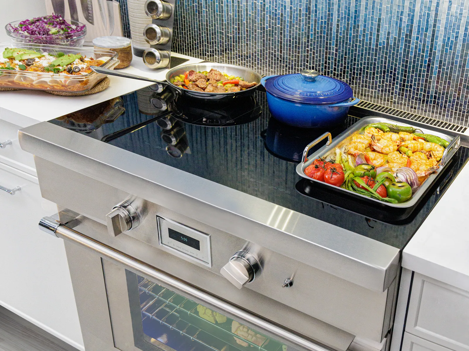 Of course! I'm happy to provide the alt text for this vibrant kitchen scene.  ***  A stainless steel induction range is showcased in a modern kitchen, its black glass cooktop filled with a variety of colorful dishes. A griddle with roasted shrimp and vegetables, a blue Dutch oven, and a frying pan demonstrate the appliance's cooking capacity, all set against a vibrant blue mosaic tile backsplash.