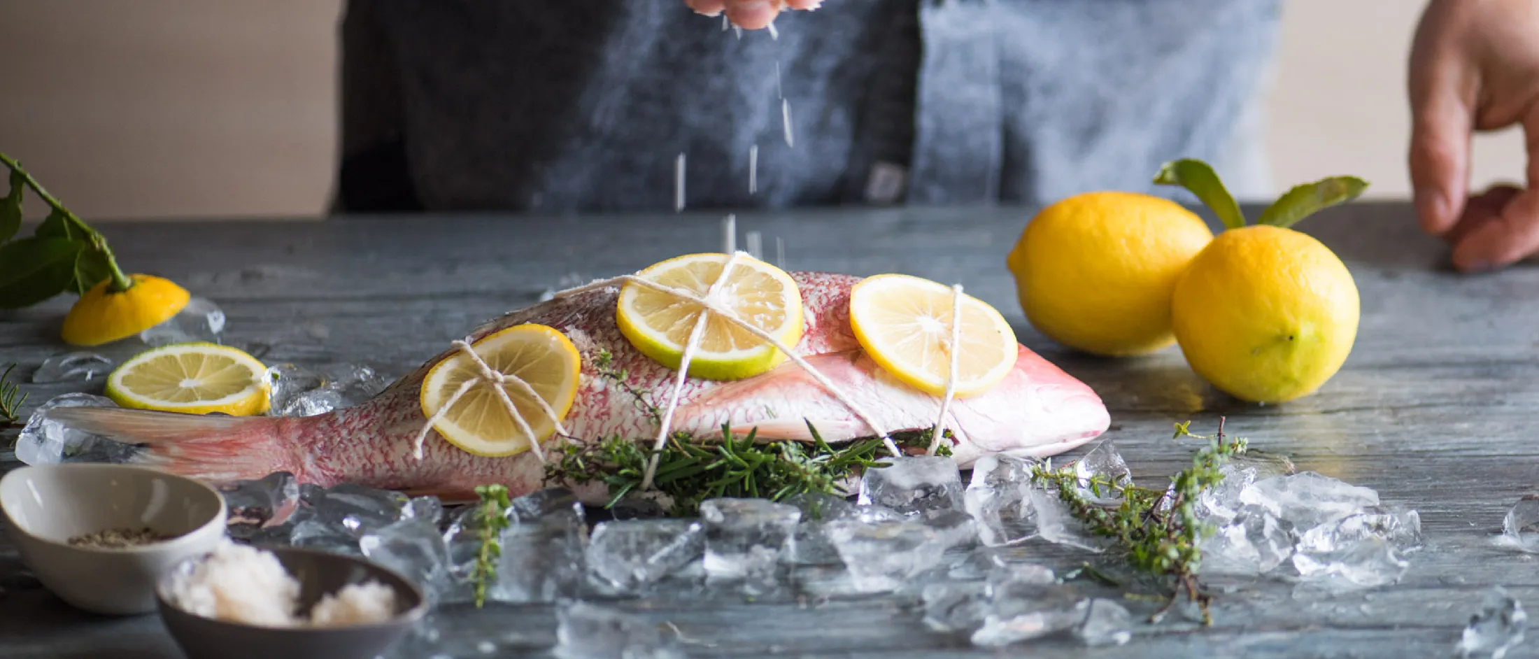 Whole fish on herbs with lemon slices tied on top surrounded by ice cubes and lemons with a person sprinkling salt over it