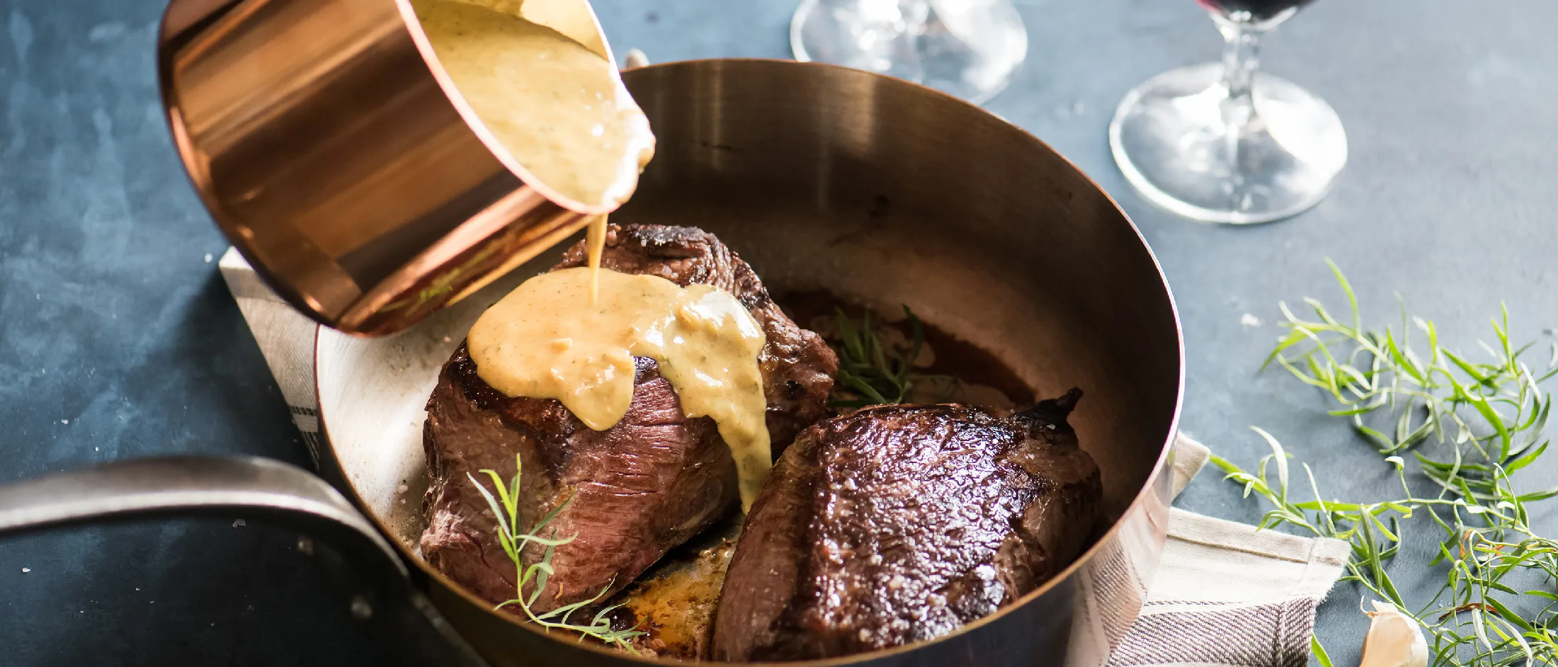 Two cooked sirloin steaks in a pan with sauce being poured over one steak and two glasses of red wine in the background