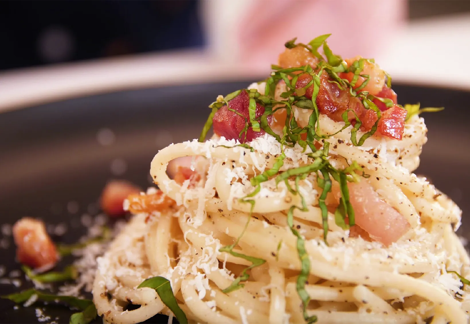 Plate of spaghetti cacio e pepe topped with grated cheese, black pepper, chopped bacon, and fresh basil strips