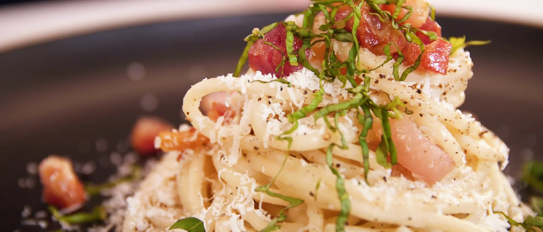 Plate of spaghetti cacio e pepe topped with grated cheese, black pepper, chopped bacon, and fresh basil strips