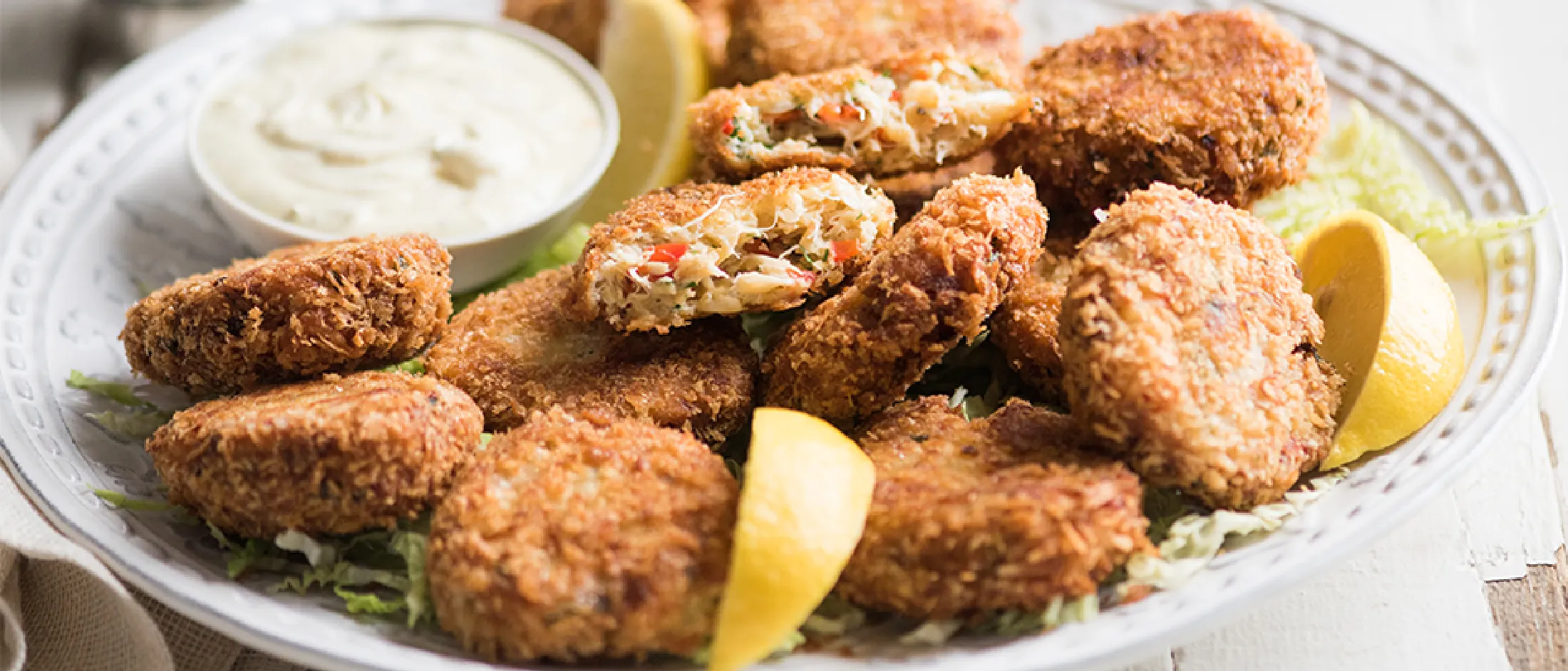 Plate of breaded crab cakes with lemon wedges and a bowl of dipping sauce on a light surface