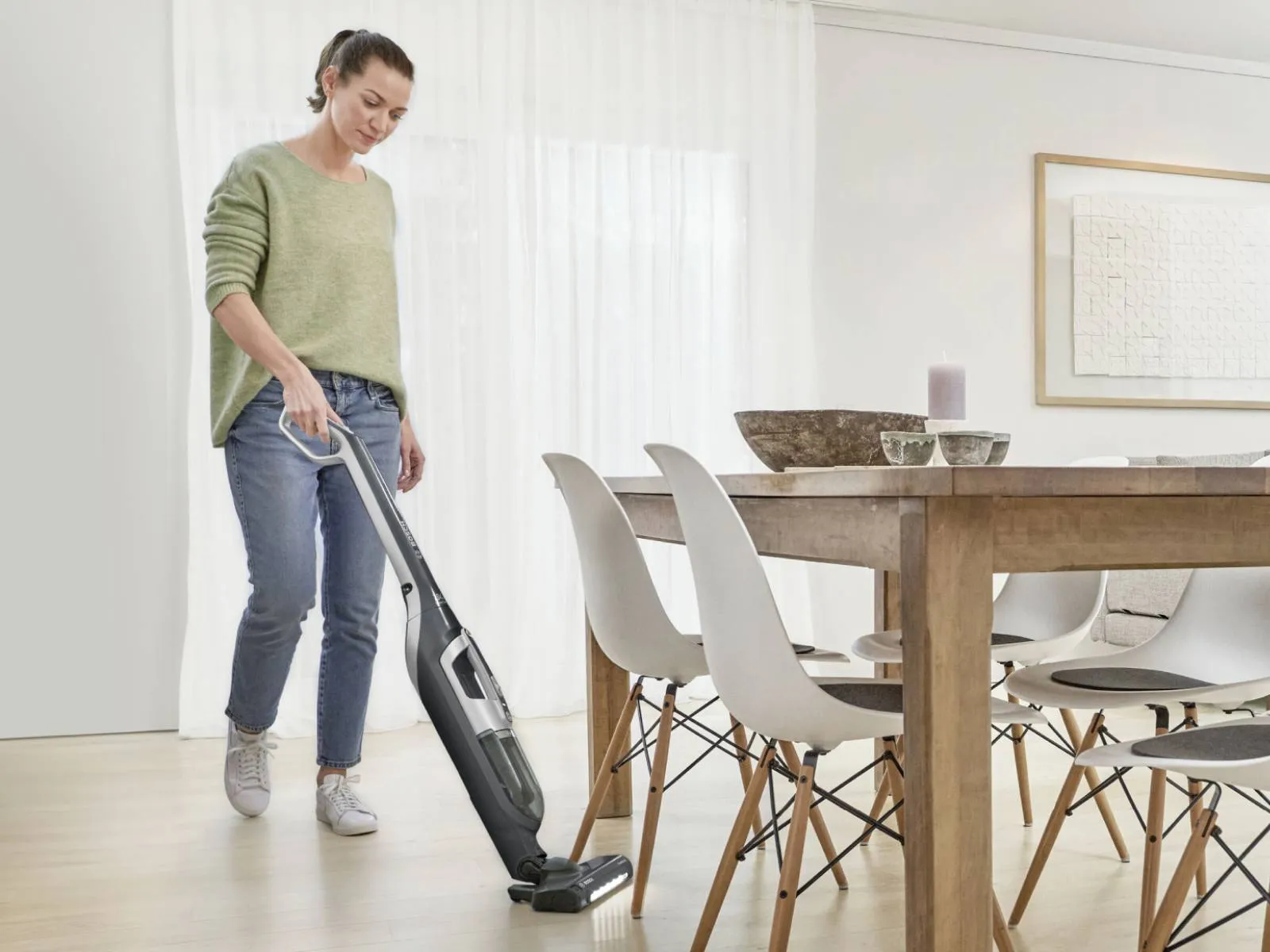 A woman vacuums a light wooden floor with a Bosch cordless stick vacuum cleaner in a bright, modern dining room.