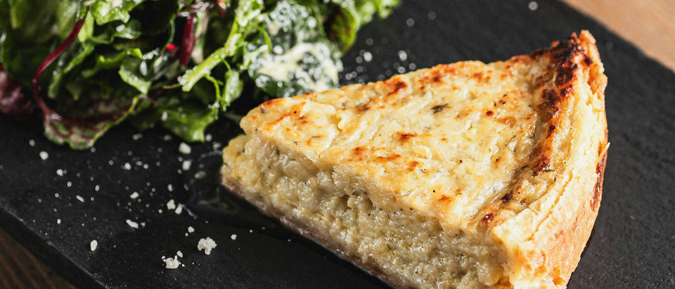 Slice of quiche with a side of leafy green salad on a dark rectangular plate