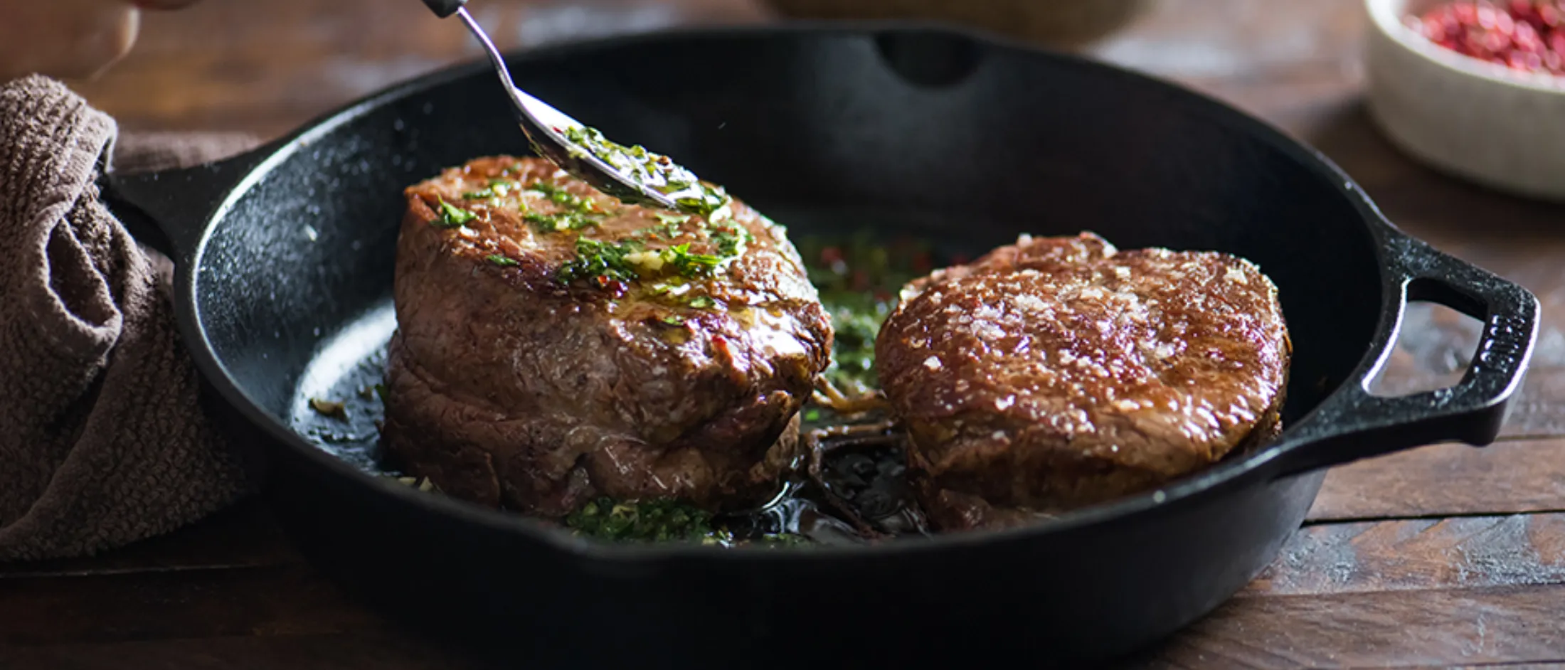 Two fillet mignon steaks cooking in a cast-iron skillet with a hand spooning green chimichurri sauce over one steak