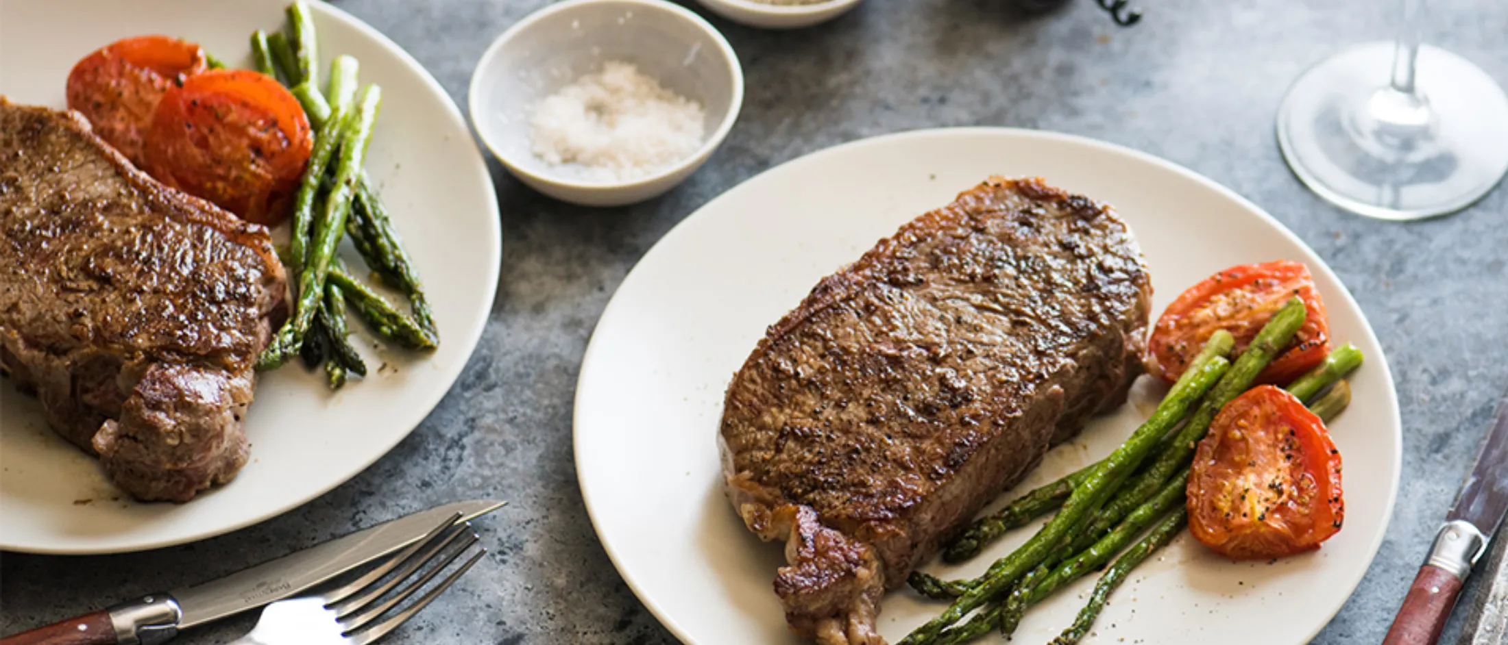 Two plates each with a cooked ribeye steak, grilled asparagus, and tomato halves on a gray surface with cutlery, wine glasses, and small bowls of salt and pepper