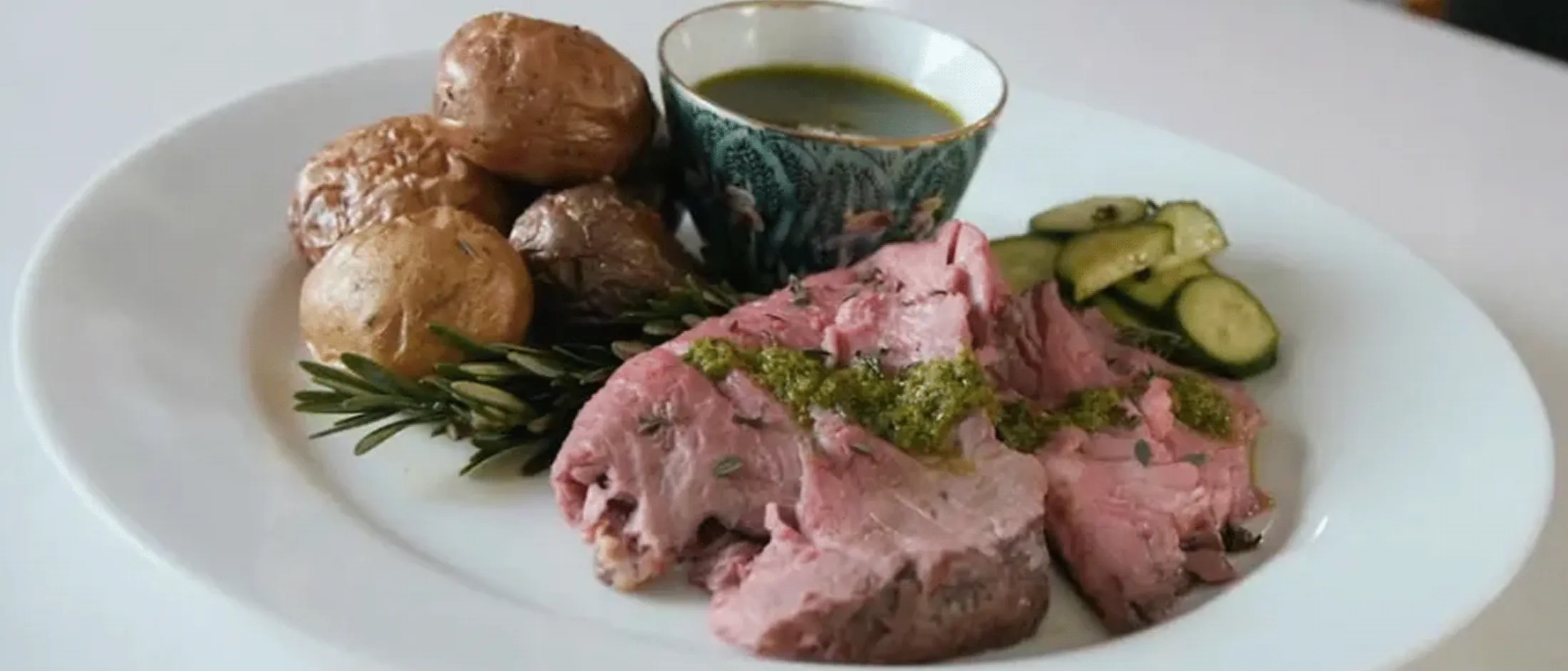 Plate with sliced leg of lamb topped with green sauce, roasted potatoes, sliced pickles, and a small bowl of green sauce