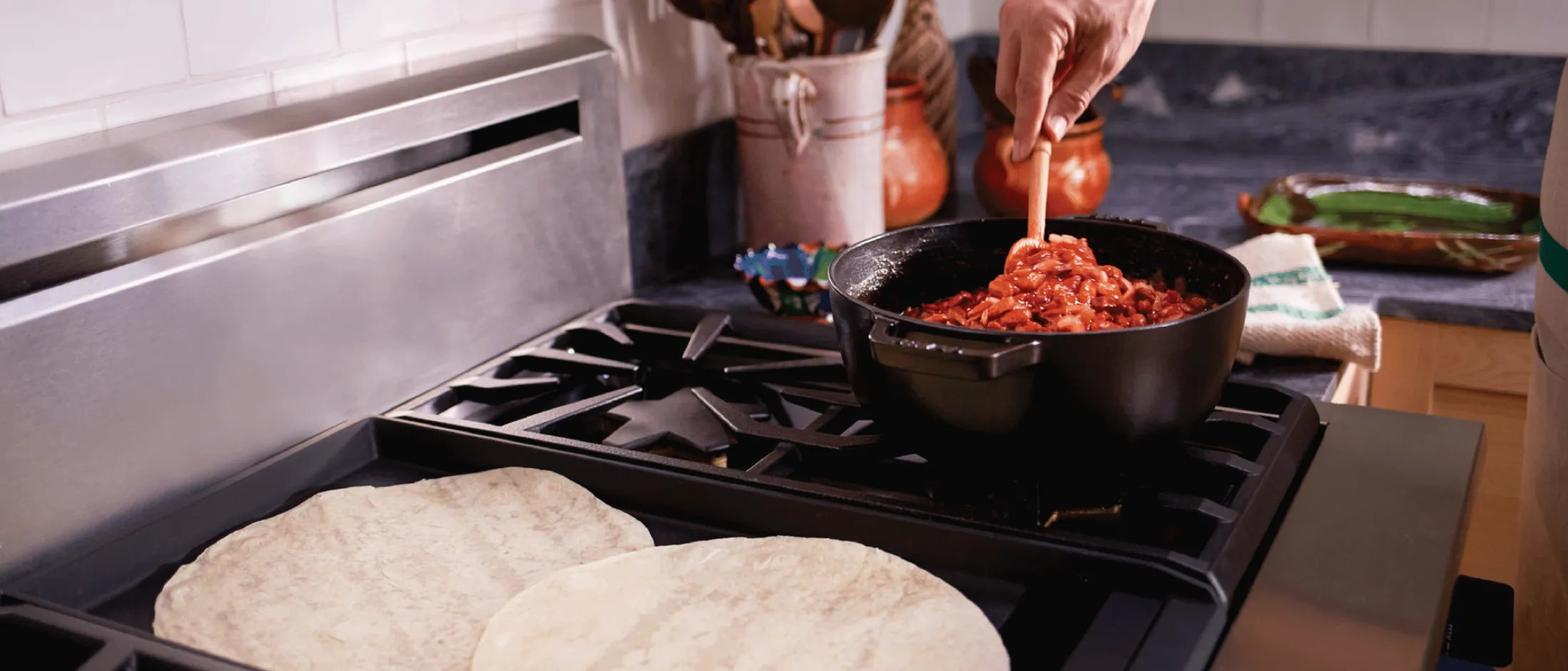 Hand stirring a pot of red sauce on a stove with two tortillas warming on the burner