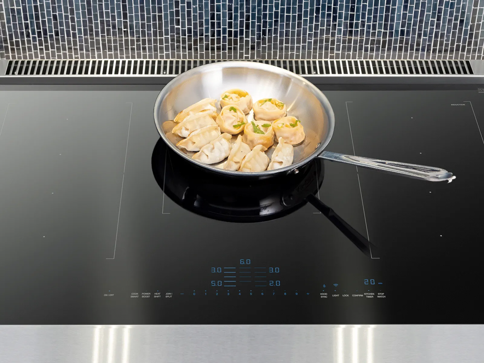 A stainless steel frying pan filled with dumplings sits on a sleek, black induction cooktop with illuminated blue digital controls, set against a modern blue mosaic tile backsplash.