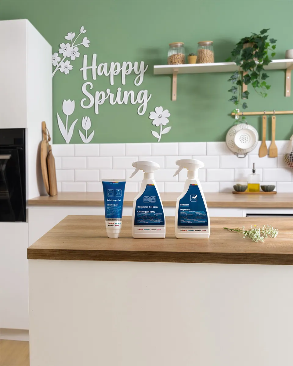 Bancada da cozinha com três produtos de limpeza, parede verde com texto "Happy Spring" e decalques florais e azulejo branco.