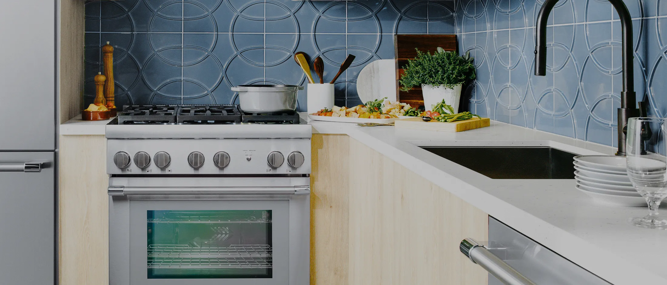 A stainless steel Thermador gas range installed in a modern kitchen with light wood cabinets and a white countertop. The kitchen features a distinctive blue circular-patterned tile backsplash, and the counter is prepped with fresh ingredients and cookware.