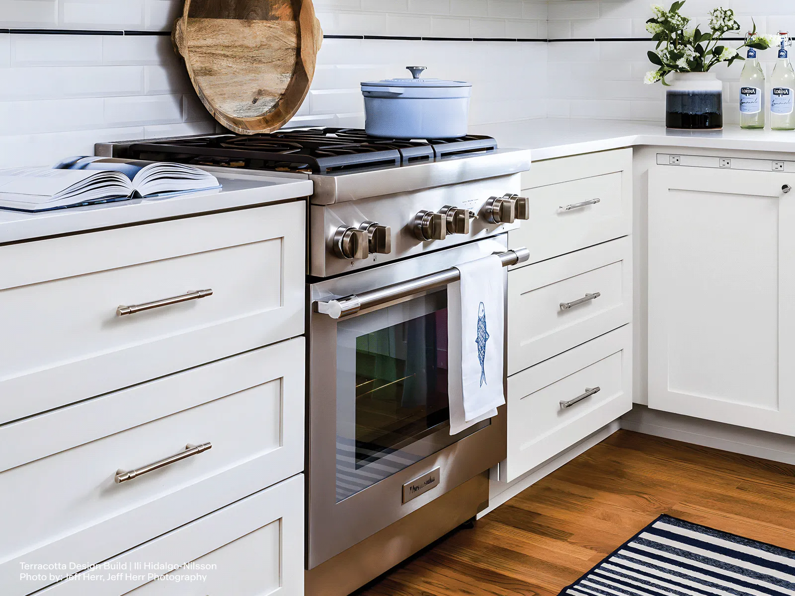 A stainless steel Thermador gas range is installed in a bright, modern kitchen with white shaker cabinets and a white countertop. A light blue Dutch oven sits on the cooktop, and a white tea towel with a blue fish illustration hangs from the oven handle. The kitchen is styled with an open cookbook, a vase of flowers, and a blue and white striped rug on the hardwood floor.