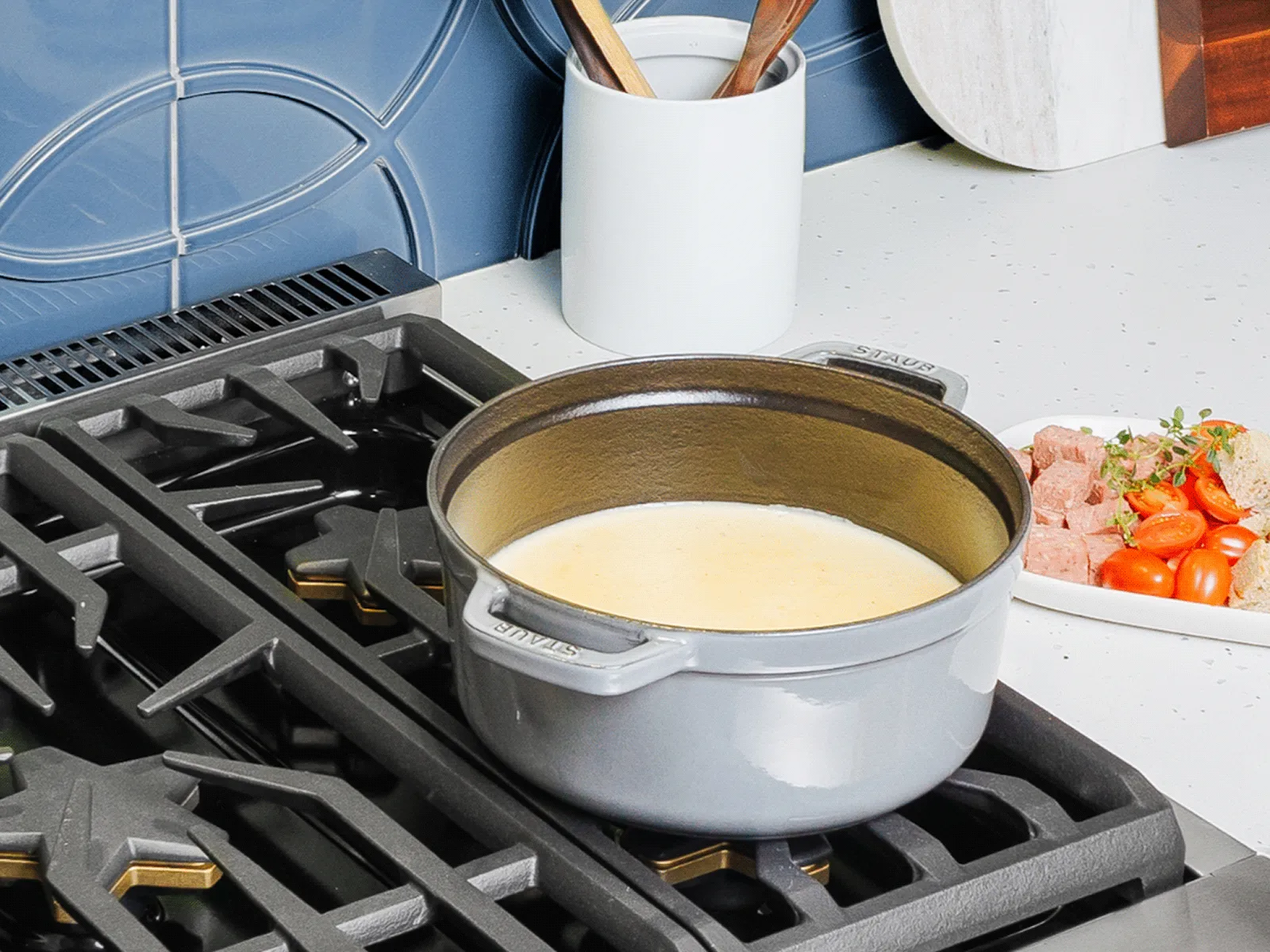 A gray Staub Dutch oven filled with creamy cheese fondue simmers on a professional-style gas cooktop with black cast-iron grates. On the white countertop beside it, a plate holds an assortment of dippers, including bread, cherry tomatoes, and cubed sausage, all set against a modern blue-tiled backsplash.
