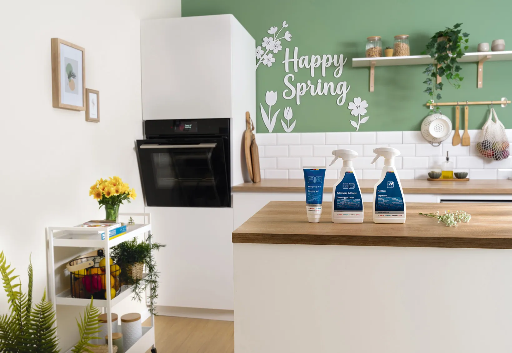 Bancada da cozinha com três produtos de limpeza, parede verde com texto "Happy Spring" e decalques florais e azulejo branco.