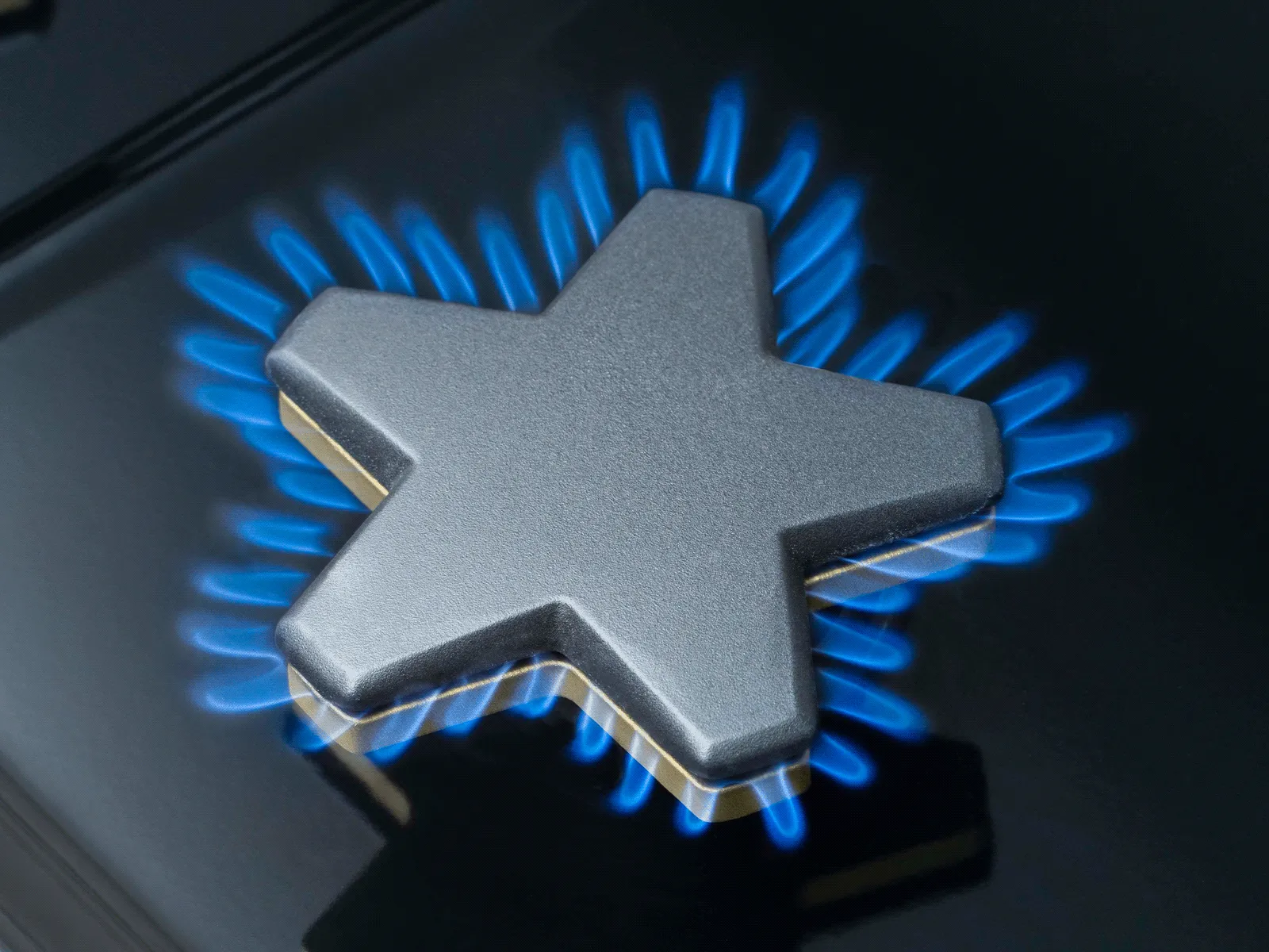 A close-up of a black gas stovetop burner with a unique star-shaped grate. Bright blue flames are visible around the edges of the star, indicating the burner is on. The surface of the stovetop is reflective.