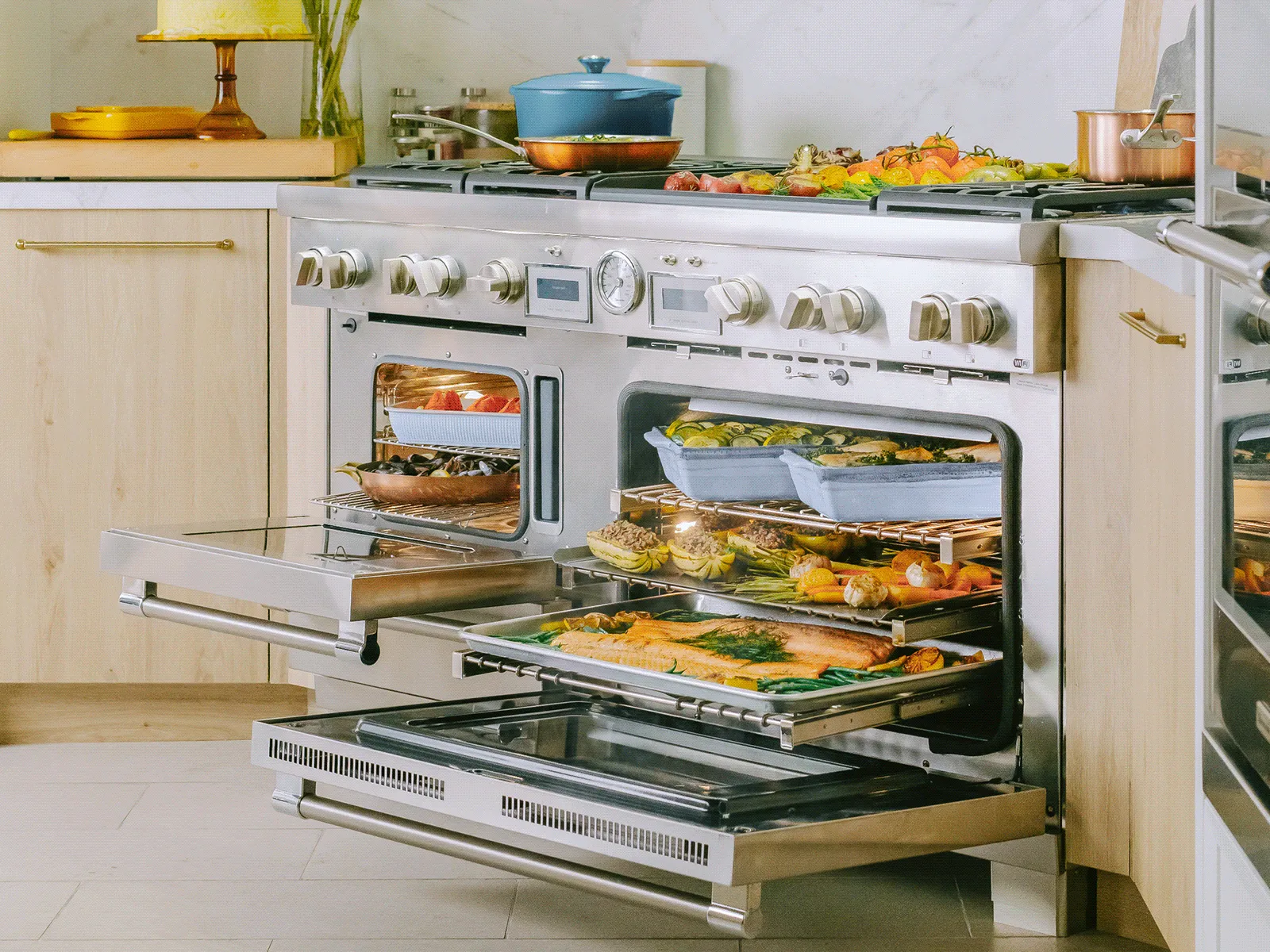 A large stainless steel range with open ovens revealing multiple dishes cooking, including fish and vegetables, with more food on the stovetop.