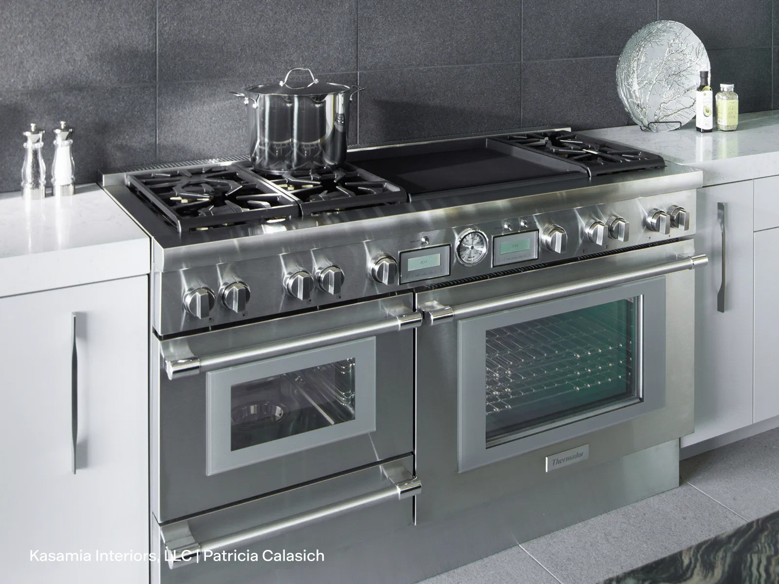 Stainless steel dual oven with six gas burners and a pot on the stove, set in a modern kitchen with white countertops and gray tile backsplash.