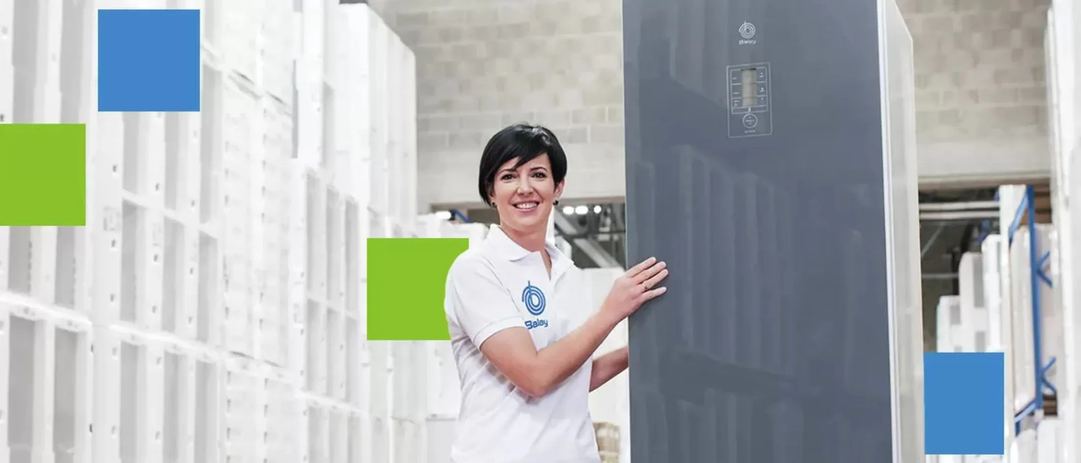 Trabalhador com um polo branco a segurar um eletrodoméstico alto e escuro, dentro de um armazém ou arrecadação com prateleiras brancas e caixas embaladas.