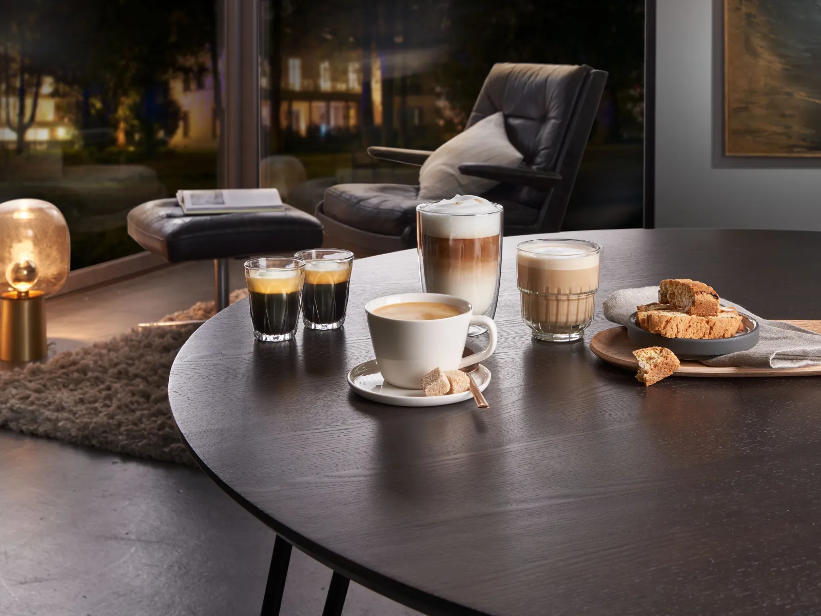 Five coffee specialities and biscuits on a dark dining table in a cosy evening living room setting.