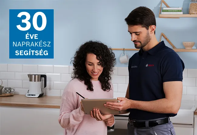 Egy Bosch-technikus kesztyűben javít egy készüléket egy modern konyhában, miközben egy nő figyelmesen nézi; szerszámok vannak a pulton mellette.
