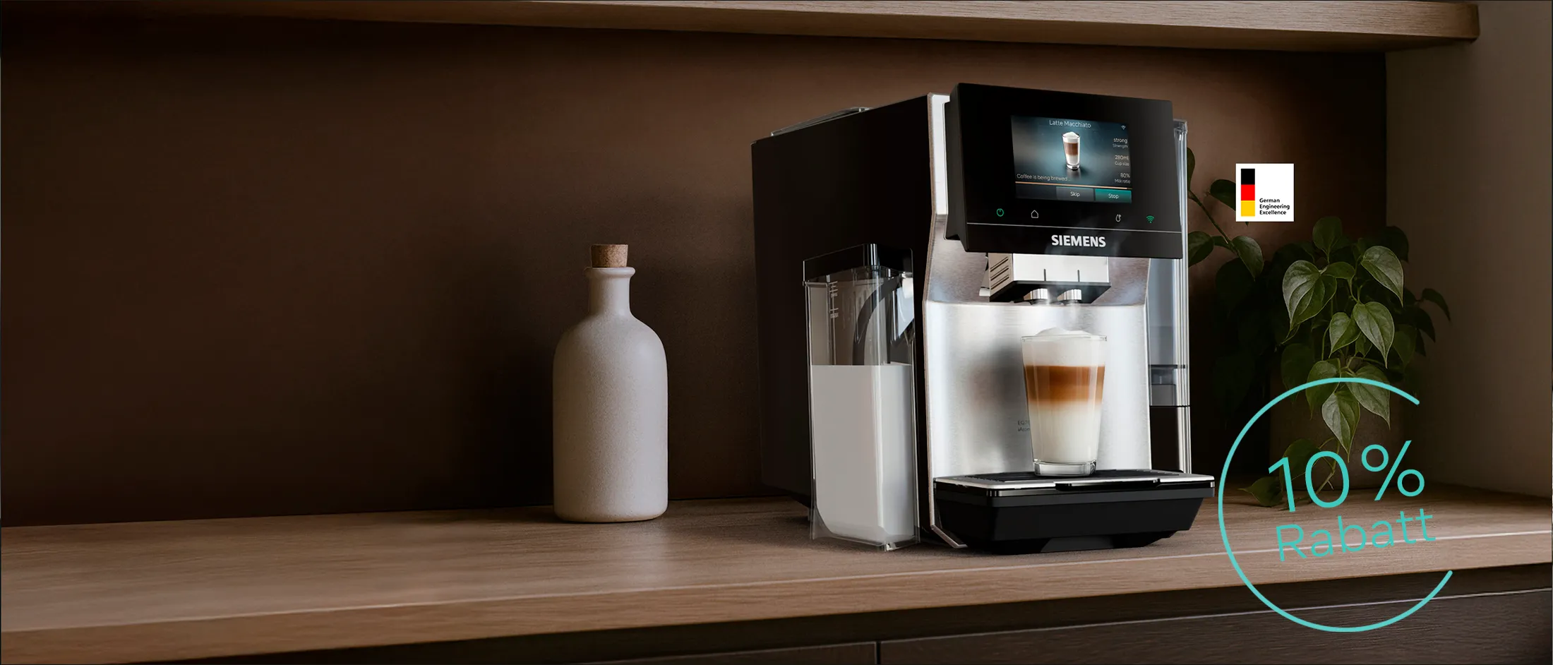 Siemens Kaffeemaschine mit angebrachtem Milchbehälter auf einer Holzarbeitsplatte mit einem geschichteten Latte Macchiato; kleine beige Vase mit Holzdeckel und kleine Pflanze in der Vase. „German Engineering Excellence“-Logo sichtbar.