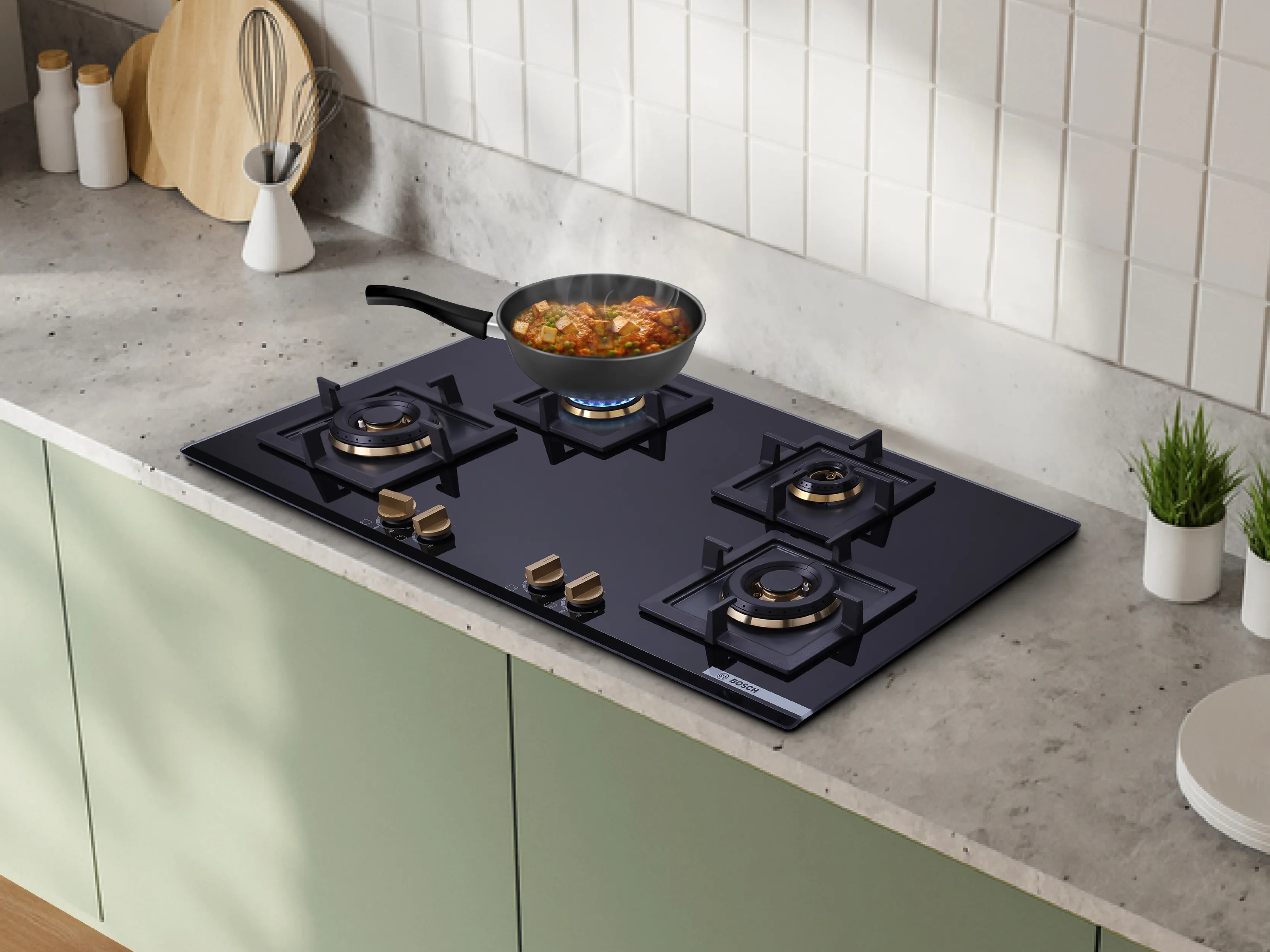 Black glass four-burner gas cooktop set into a gray countertop with a steaming skillet on a lit burner, brass knobs, utensils, and small potted plants nearby.