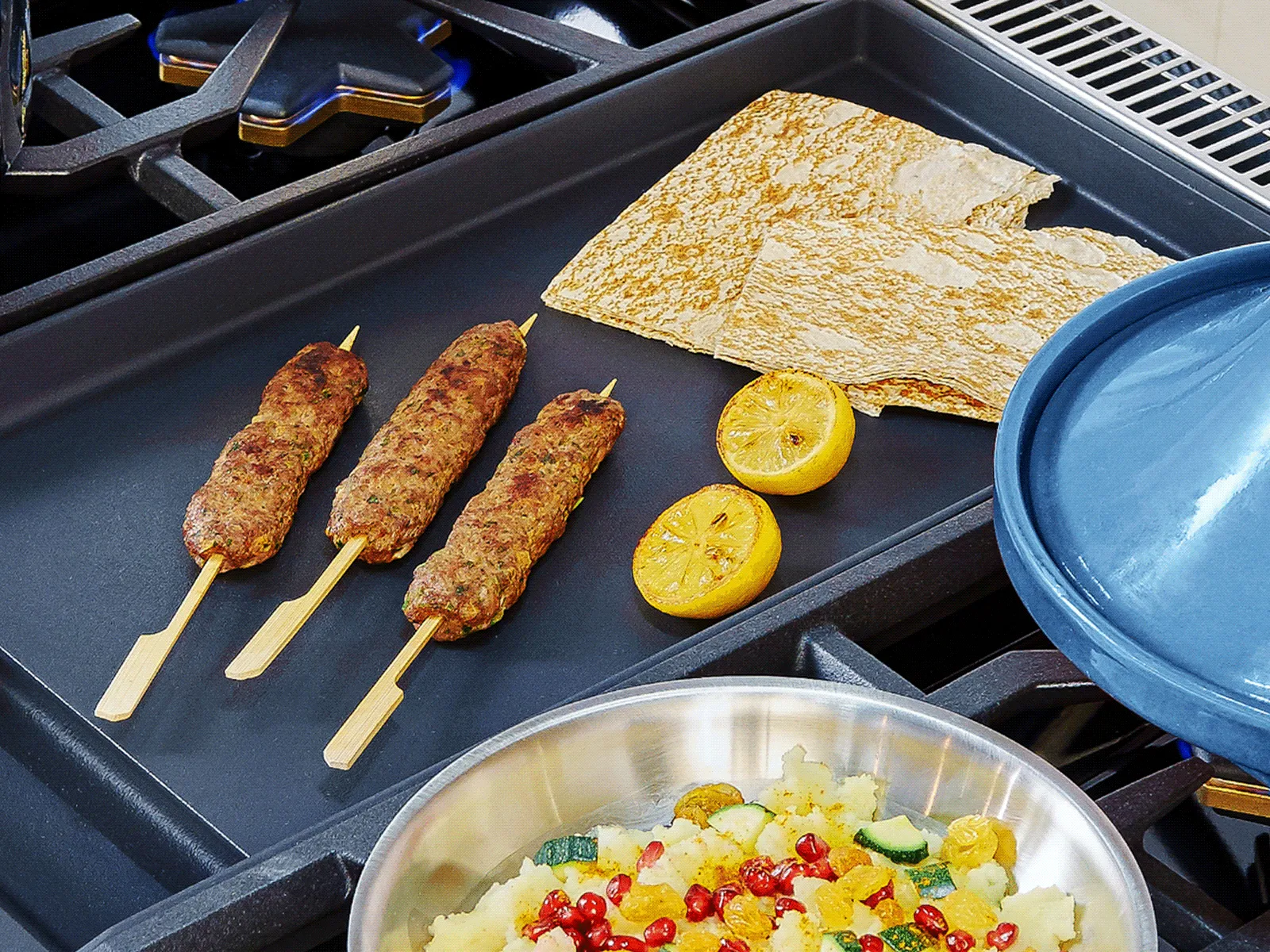Three grilled meat skewers, flatbreads, and halved grilled lemons on a griddle next to a pan with colorful cooked vegetables.