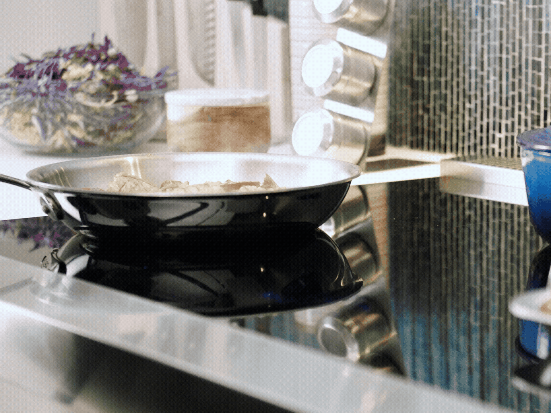A person moves various cookware, including a blue Dutch oven and a sheet pan of shrimp and vegetables, across the surface of a black induction cooktop.