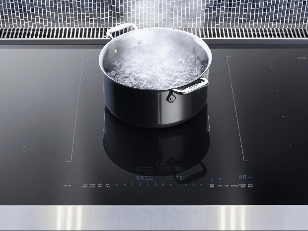 A close-up of a stainless steel pot of water at a rolling boil on a sleek, black induction cooktop with illuminated blue digital controls.