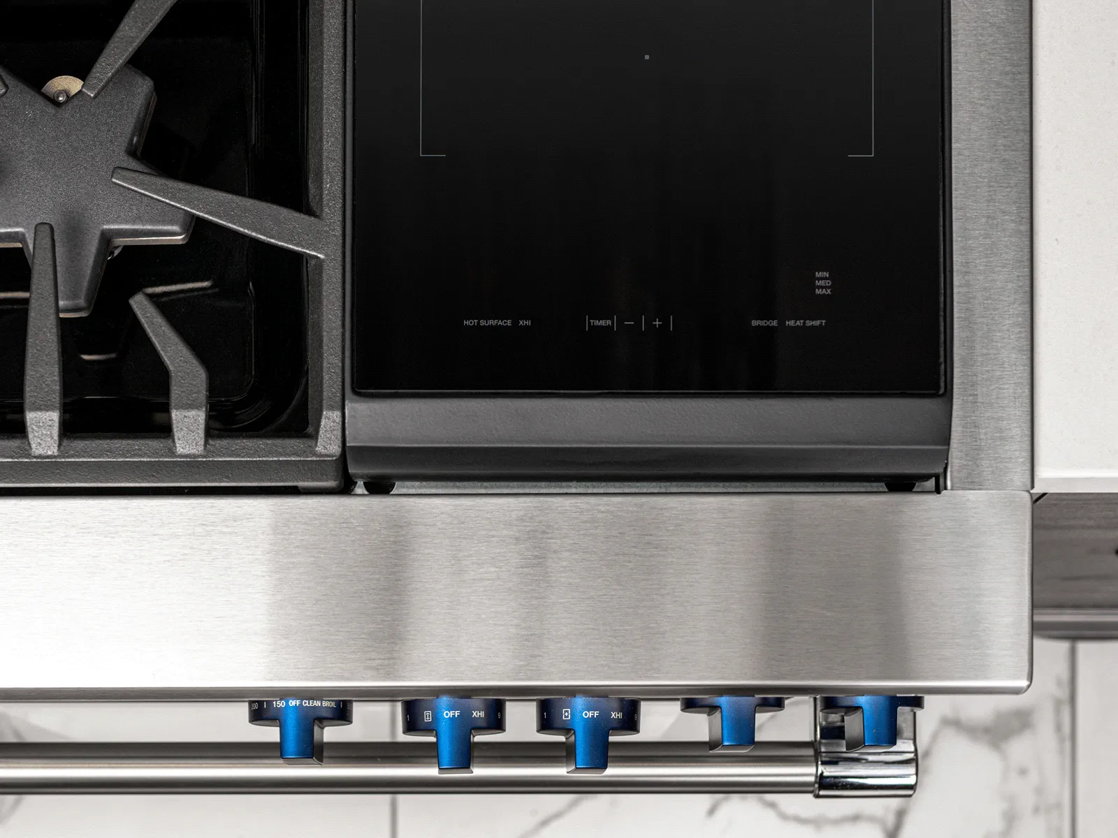 A close-up, overhead shot of a professional-style range, highlighting a cooktop that combines a star-shaped gas burner on the left with a sleek, black glass induction zone on the right. The stainless steel front features a row of signature blue metallic control knobs.