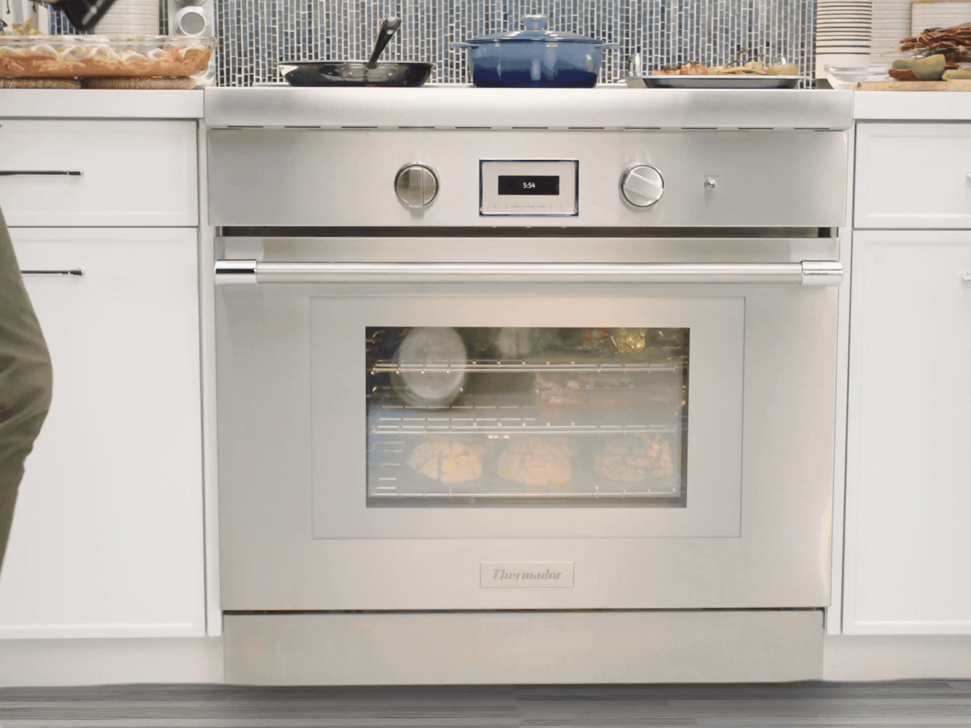 A person opens a stainless steel Thermador oven, revealing three fully loaded racks with various dishes, including colorful concha pastries, a casserole, and stuffed peppers.