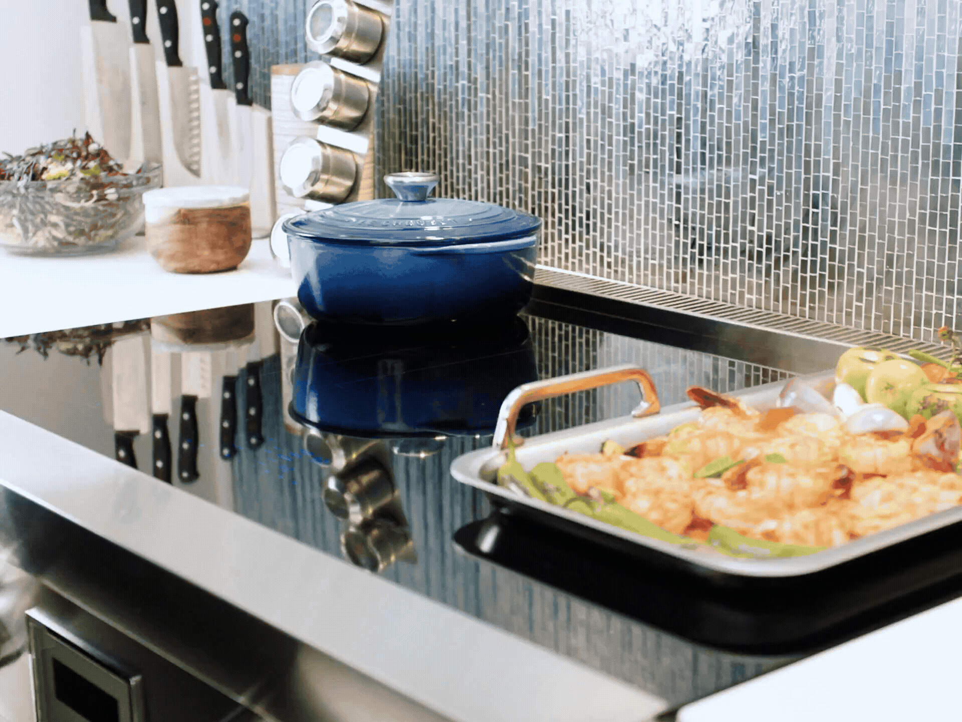 A person moves various cookware, including a blue Dutch oven and a sheet pan of shrimp and vegetables, across the surface of a black induction cooktop.