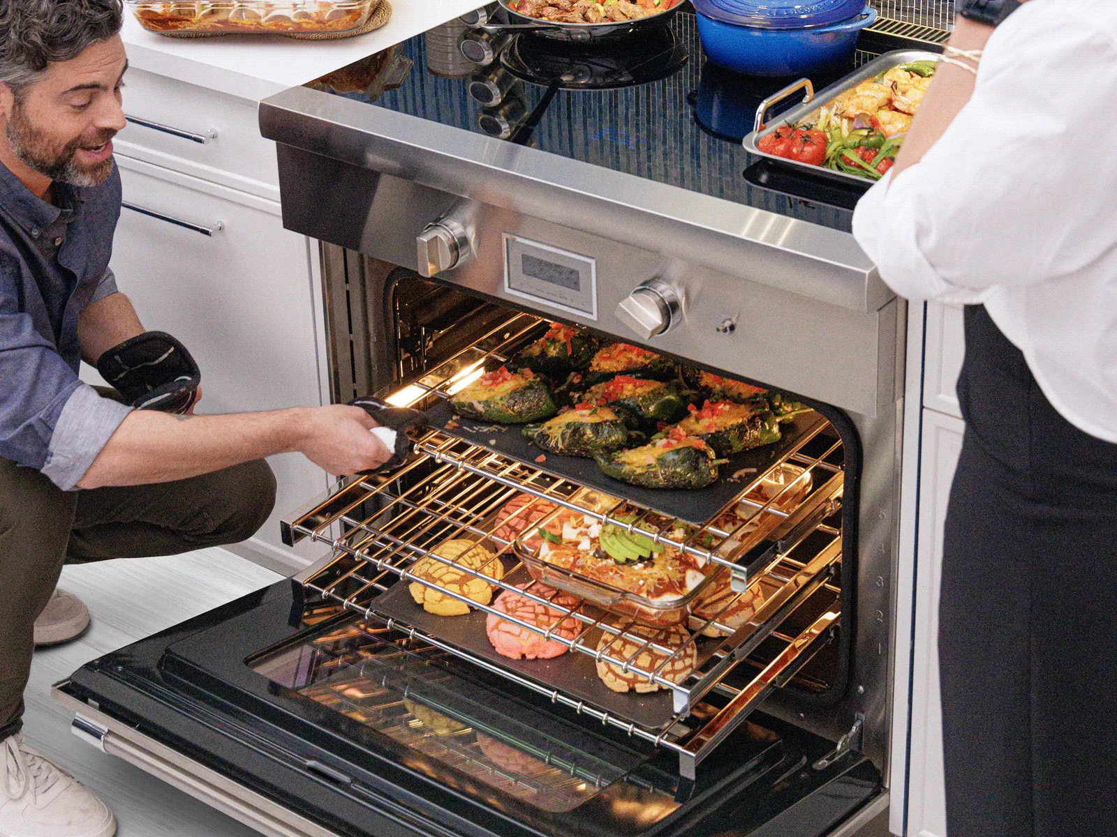 A smiling man crouches in a modern kitchen, pulling a rack of stuffed peppers from a large, open stainless steel oven. The oven is filled with food on multiple racks, including colorful concha pastries and a casserole, while a woman stands nearby at the cooktop.