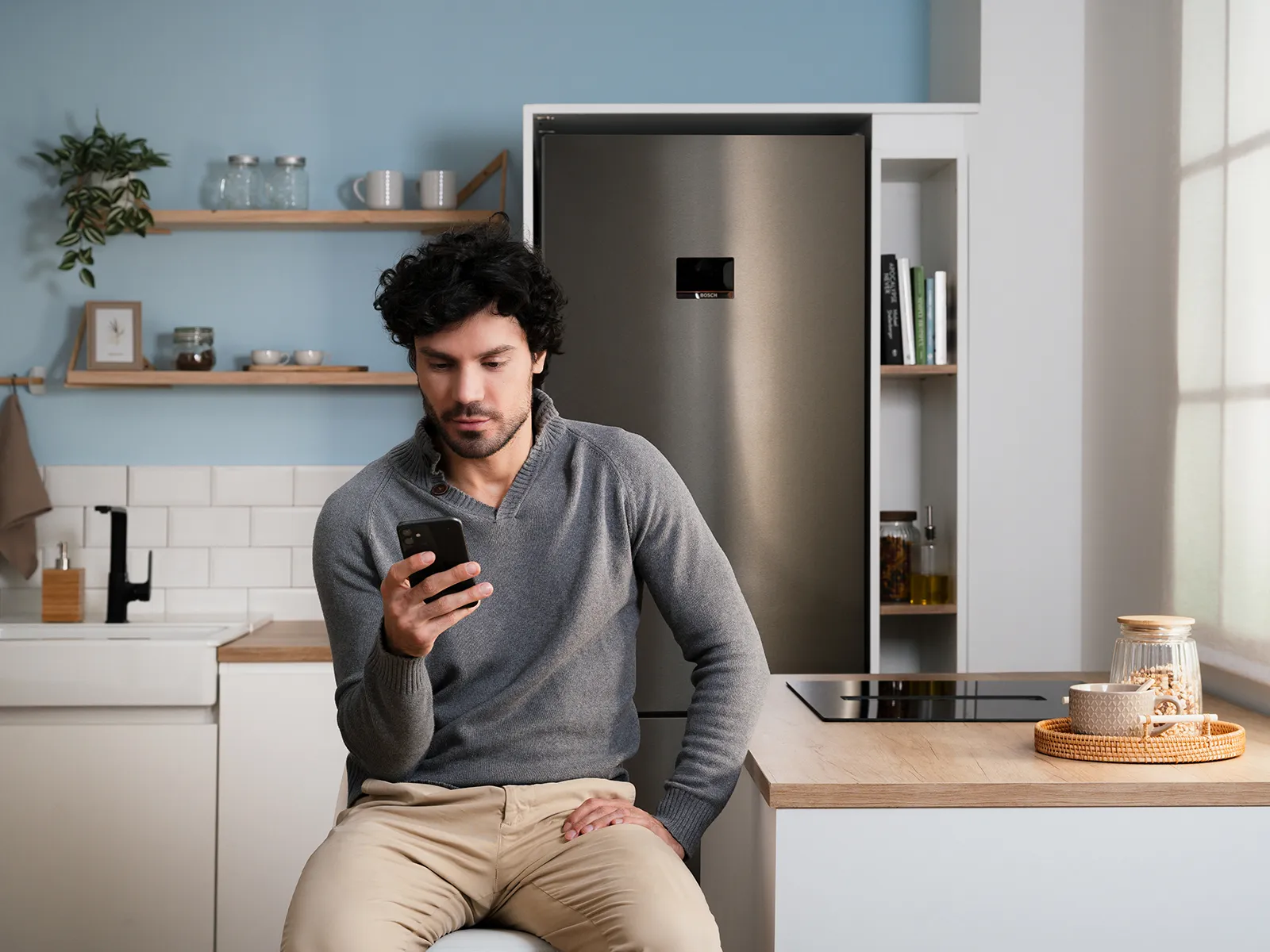 Person sitzt auf einer modernen Küchentheke, trägt einen grauen Pullover und hält ein Telefon; Edelstahl-Kühlschrank von Bosch, offene Regale, Spüle und Kochfeld.