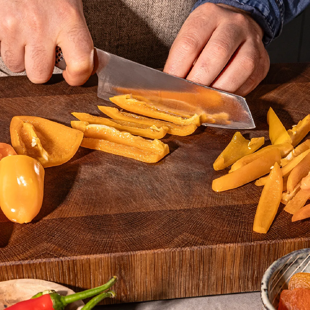 Person slicing orange bell pepper into strips on a wooden cutting board with other peppers and dried apricots nearby.